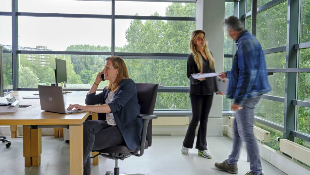 Een vrouw zit achter haar laptop te bellen terwijl er twee mensen achter haar aan het praten zijn