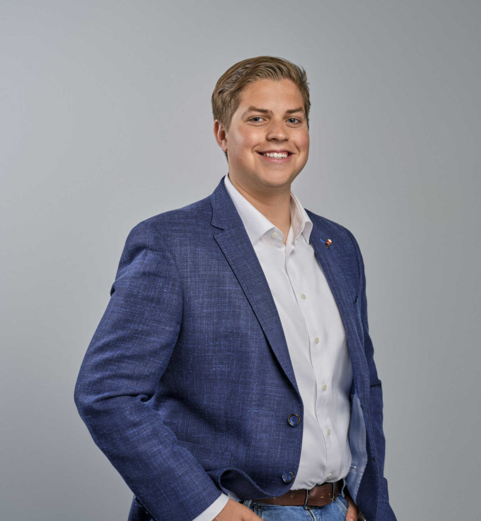 Cedric: Man in een wit overhemd en een donkerblauw colbert die lacht en zijn handen in zijn zak heeft.
