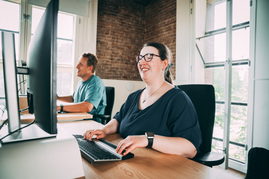 Een man en een vrouw zijn achter de computer aan het werk. De vrouw gebruikt hulpmiddelen.