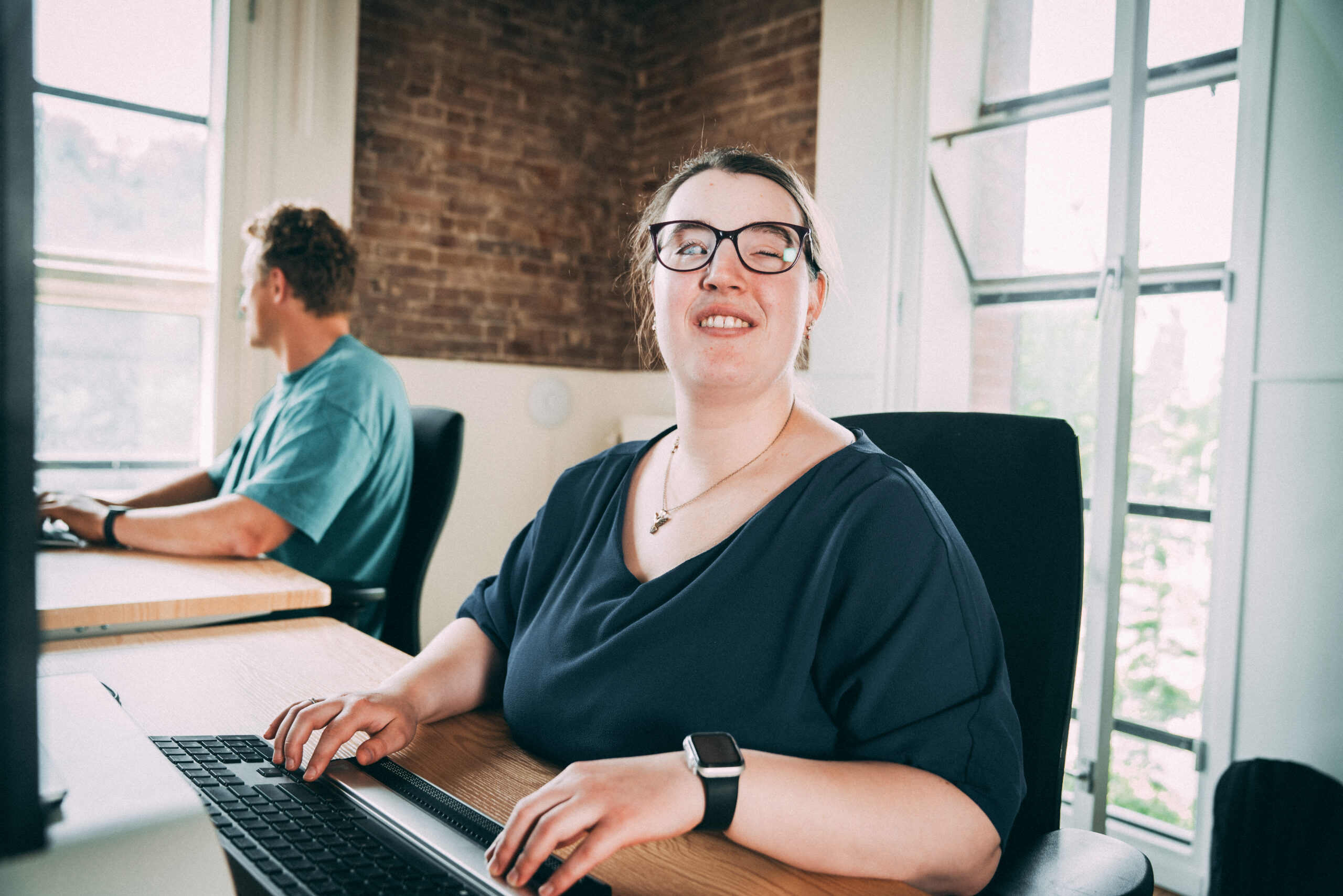 Een vrouw zit achter haar computer te werken met haar brailletoetsenbord. Ze kijkt naar de camera en lacht.
